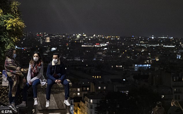 People wear face masks as they look at the Paris skyline from Montmartre at nightfall, just hours before a city-wide curfew goes into effect in Paris, France